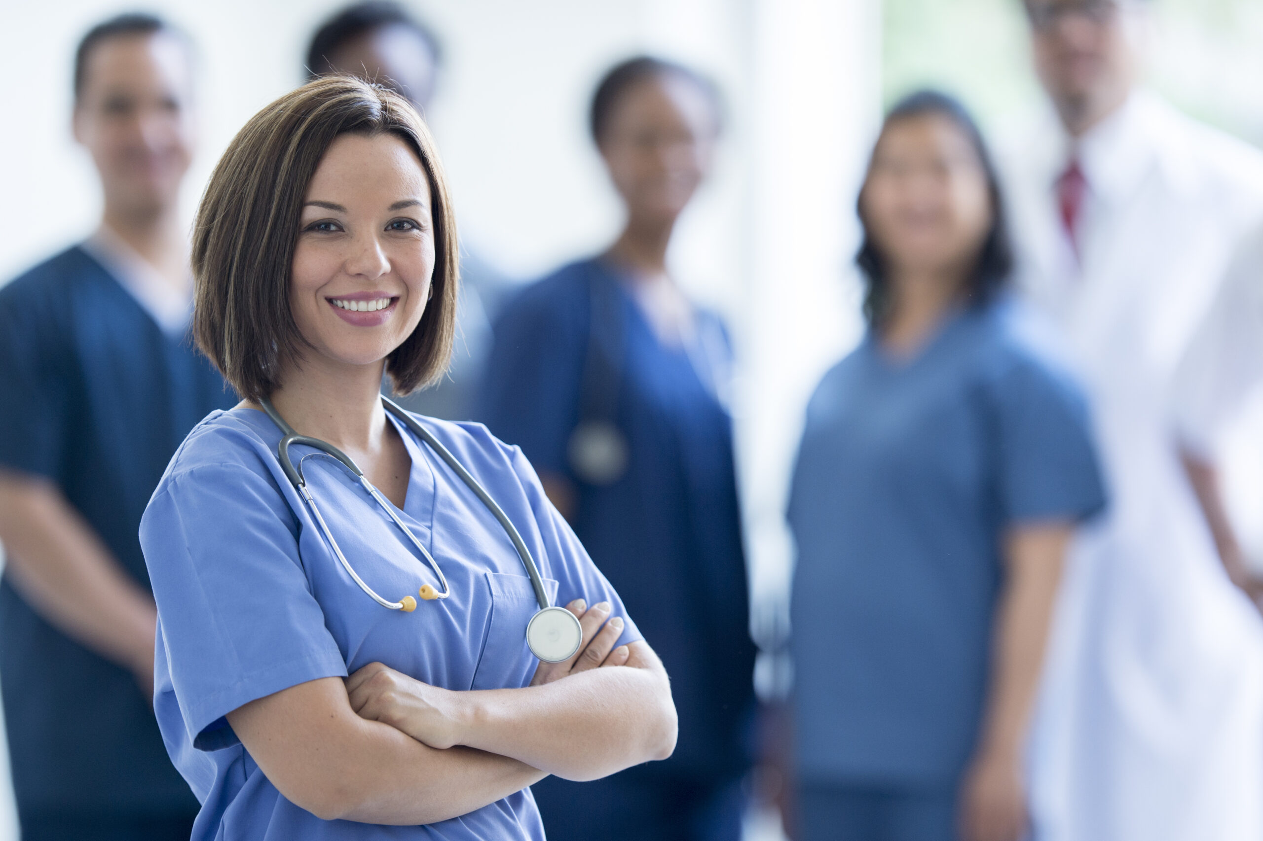 A group of healthcare professionals, including a smiling female nurse in the foreground wearing blue scrubs and a stethoscope, with four other medical staff members standing behind her in a well-lit clinical setting.